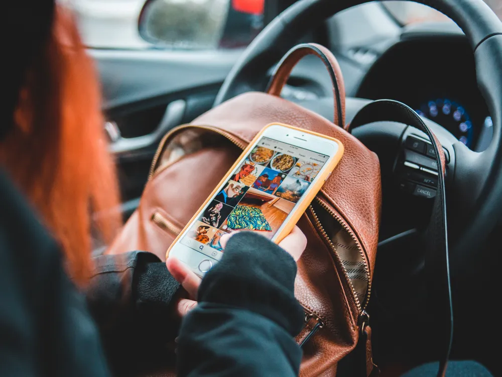 A person with red hair holds a smartphone displaying colorful Instagram photos while sitting in a car, with a brown bag nearby.