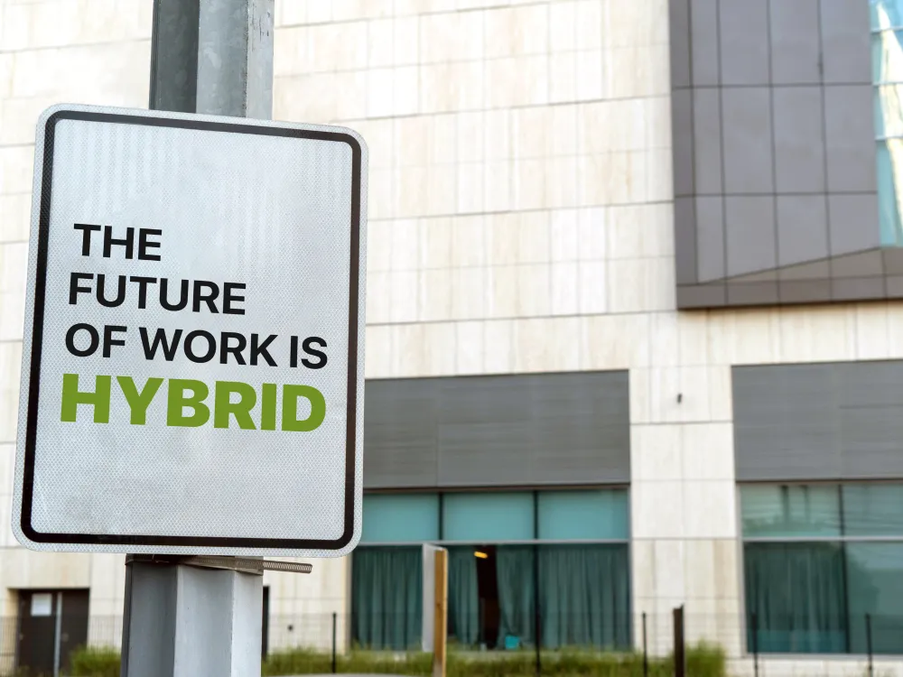 A sign reads, "The future of work is HYBRID," in front of a modern building with glass windows and beige walls.