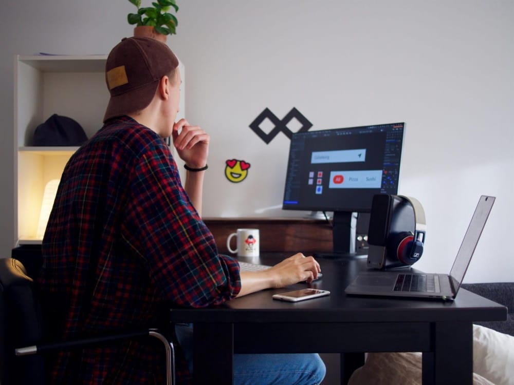 A person in a plaid shirt sits at a desk, focused on a computer screen displaying a search interface, with headphones and a phone nearby.