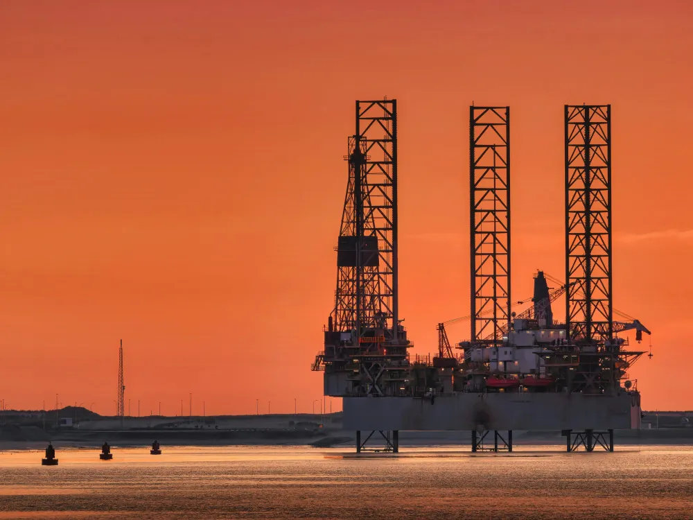 Silhouette of a drilling rig against a vibrant orange sunset, with calm waters and distant boats.