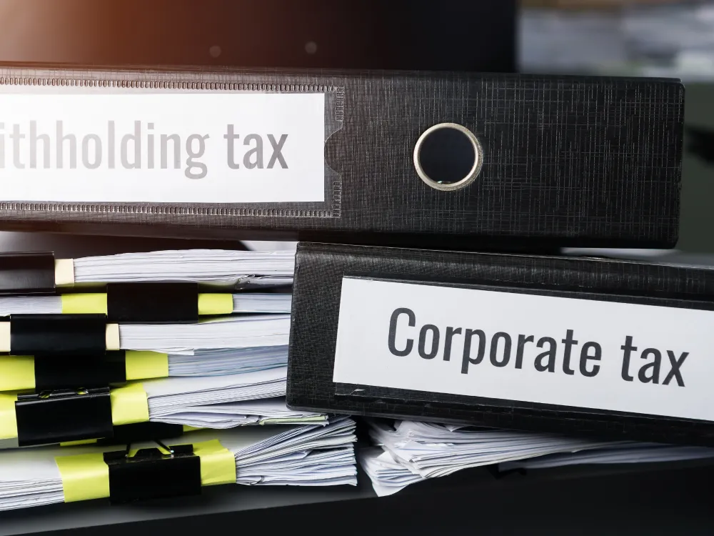 Black office binders labeled “Withholding tax” and “Corporate tax” stacked on top of paperwork files with sticky notes, symbolizing business taxation and financial administration.