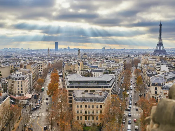 A panoramic view of Paris featuring the Eiffel Tower, city streets, and rays of sunlight breaking through clouds. Autumn foliage is visible.