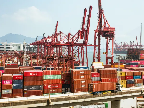 A busy shipping port filled with colorful containers and towering red cranes against a backdrop of the sea and distant mountains.