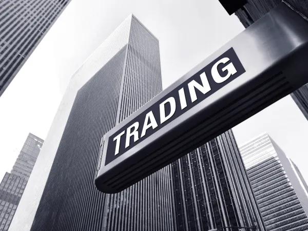 A close-up view of a sign reading "TRADING" against a backdrop of tall, modern skyscrapers in a black and white setting.
