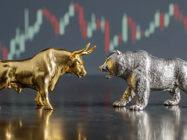 Gold bull and silver bear statues facing each other on a reflective surface, with blurred red and green stock market chart bars in the background.